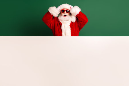 Santa in red suit with white beard posing behind a large blank banner on a green background celebrating the holiday season and Christmas vibesの写真素材