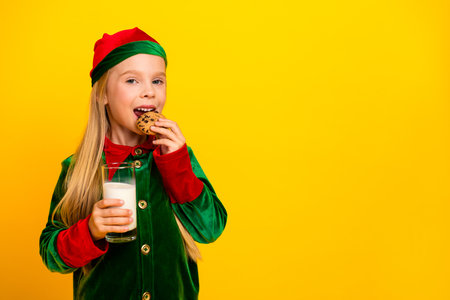 Adorable child in elf costume enjoys festive snack holding milk and cookie on bright yellow background, spreading Christmas joyの写真素材