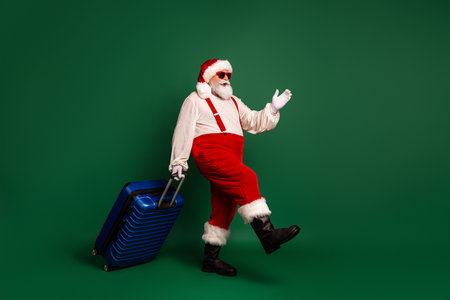 Santa in red suit with blue rolling suitcase on a green studio backdrop ready for holiday travel and Christmas marketingの写真素材