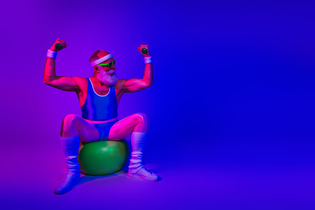 Bearded senior man in retro fitness outfit strikes a powerful pose on an exercise ball under neon purple and blue lights showing strength and joyの写真素材