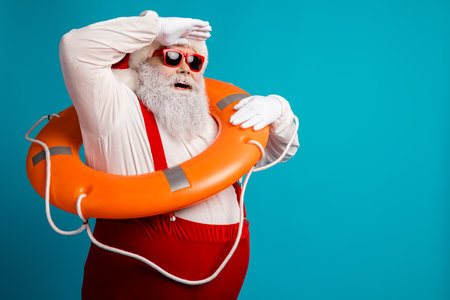 Santa Claus in red suspenders and hat holds an orange lifebuoy against a bright blue background for a playful holiday themed fashion and lifestyle stock imageの写真素材