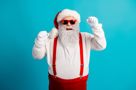 Santa in red suspenders and a white suit dances with joy on a bright blue studio background perfect for Christmas holiday campaignsの写真素材