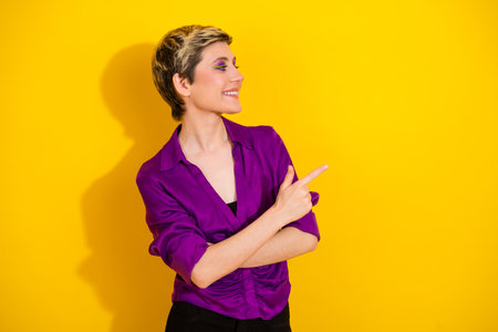 Young stylish woman in a purple blouse smiles and points against a bright yellow background for...