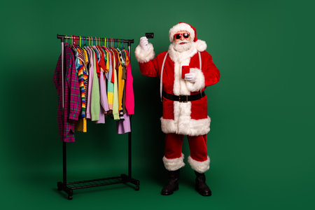 Santa poses with colorful clothes rack holding phone and card in festive fashion on green backgroundの写真素材