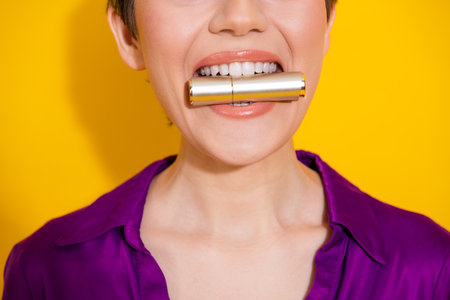Young woman in purple blouse bites a gold lipstick tube against a bright yellow background for glamour fashion advertising and beauty conceptの写真素材
