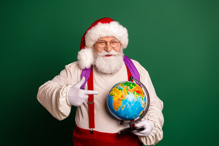 Santa Claus as grandpa traveler holding a globe during christmas season in a studio with green backgroundの写真素材