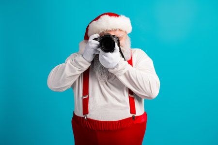 Santa photographer with camera wears red suspenders and hat against a bright blue background capturing festive moments for Christmas campaignsの写真素材