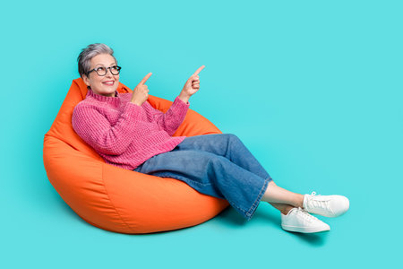Full size photo of nice person dressed knit jumper sit on pouf look directing at promo empty space isolated on turquoise color backgroundの写真素材