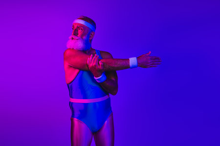 Retro fitness senior man in blue jumpsuit stretches under purple and blue neon lights in a bold studioの写真素材
