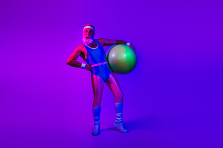 Man in blue retro athletic suit poses with large fitness ball under neon purple and blue lights in a vibrant studioの写真素材