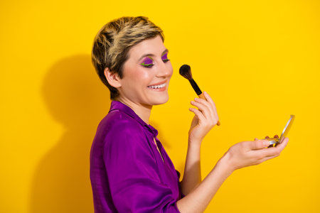 Young woman applying makeup with a smile using a compact mirror against a bright yellow background in a chic purple blouseの写真素材