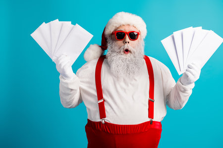 Santa in red costume with suspenders and hat holds papers against a blue background creating festive holiday vibes and Christmas shopping energyの写真素材
