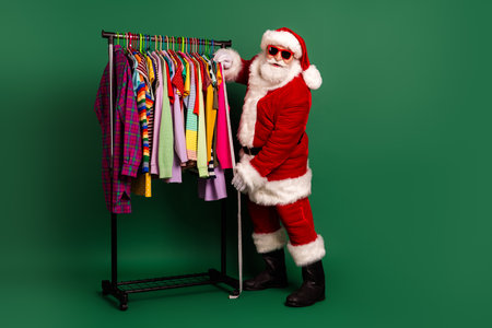 Santa in red suit with white fur posing beside a colorful clothing rack full of shirts jackets and dressesの写真素材