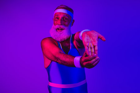 Energetic senior man in retro blue purple workout suit points forward with confident smile in neon studioの写真素材