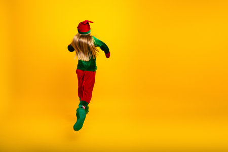 Little girl in elf costume dancing joyfully against a vibrant yellow backdrop, celebrating Christmas spirit with energyの写真素材