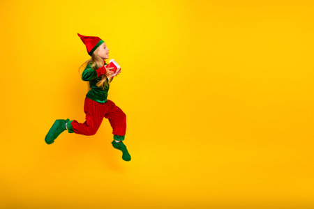 Cheerful elf kid jumping while carrying a gift against bright yellow background during festive Christmas seasonの写真素材