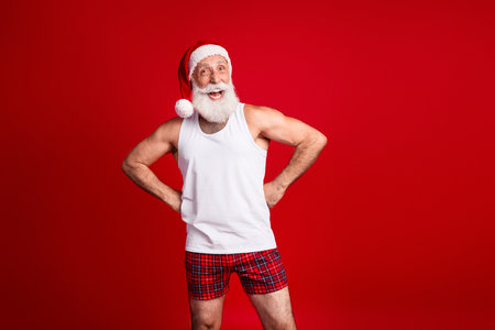 Santa Claus in white tank top with red plaid pajama shorts smiles with hands on hips in a bright Christmas studio against a bold red backgroundの写真素材