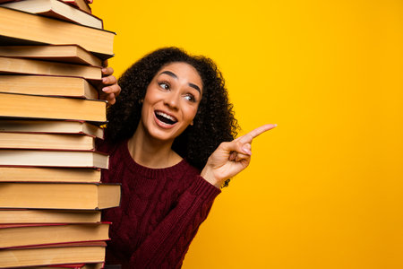 Young mixed race woman smiles and points beside a tall stack of book against a bright yellow background for education themed stock photoの写真素材