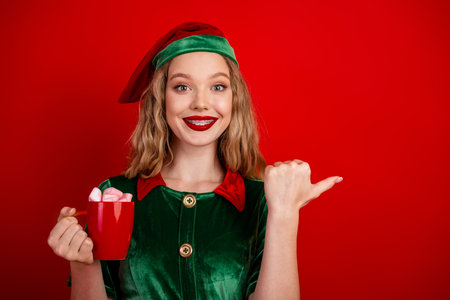 Young woman dressed as a cheerful Christmas elf holding a red mug of hot chocolate against a festive red backgroundの写真素材