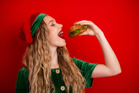 Festive young woman in elf costume enjoying a delicious burger against a vibrant red background during the holiday seasonの写真素材