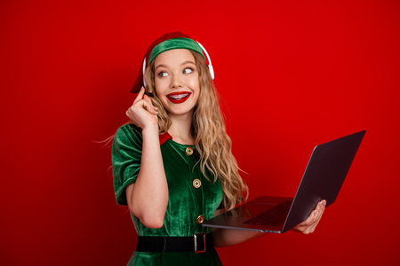 Festive holiday elf in traditional costume chatting online with laptop against red background, embodying Christmas spiritの写真素材