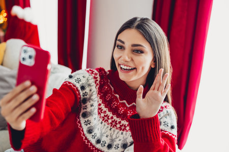 Playful holiday moment as a smiling woman in red Christmas sweater chats on her phone at home and waves hello during a festive seasonの写真素材