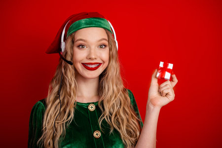 Cheerful woman dressed as an elf holding a gift against a vibrant red background during festive...