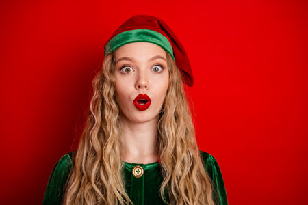 Festive blonde girl wearing a cheerful green elf costume and red hat, expressing delightful surprise against a vibrant red backgroundの写真素材