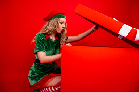 Excited young woman in Christmas elf outfit opening oversized red gift box in festive seasonal...