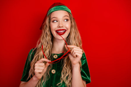 Festive young woman in green elf costume holding sunglasses against vibrant red background celebrating Christmas magicの写真素材
