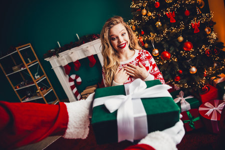 Fun festive moment as a blonde woman in a cozy sweater receives a wrapped gift by a twinkling Christmas tree in a warm home sceneの写真素材