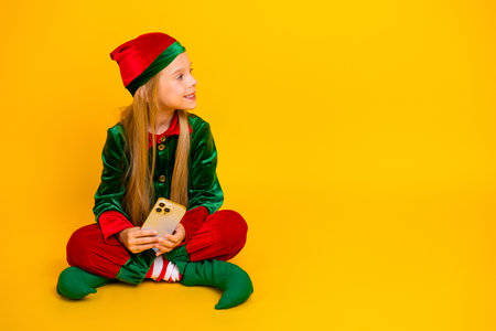 Cheerful child dressed as a Christmas elf on a vibrant yellow background holding a phone and looking joyfulの写真素材