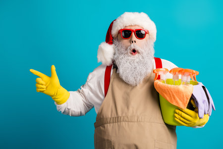 Santa in red glasses with a big white beard wearing an apron and suspenders holds cleaning supplies against a blue background for a festive holiday sceneの写真素材