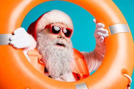 Santa with red hat and white beard wears a life jacket and sunglass while holding orange lifebuoy...