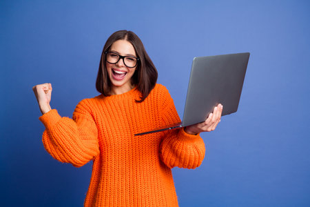 Young cheerful woman with a laptop celebrating work success wearing orange sweater standing against a blue backgroundの写真素材