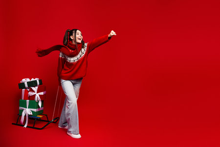 Girl in red sweater with gifts on a sleigh celebrates christmas season with joy and style against a bright red backgroundの写真素材