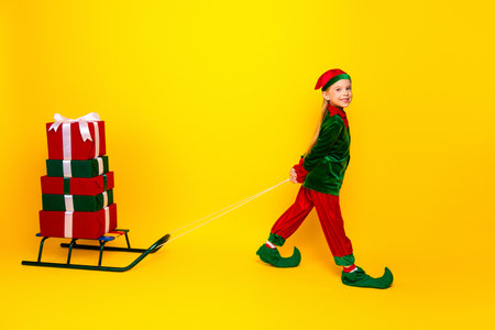 Adorable little elf pulling a sled with stacked Christmas gifts on a vibrant yellow background, bringing festive cheerの写真素材