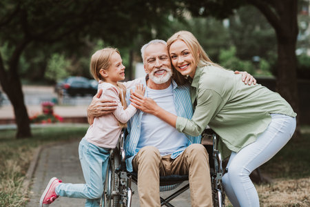 Joyful family embrace in park as grandpa in wheelchair shares love care and laughter with daughter and granddaughterの写真素材
