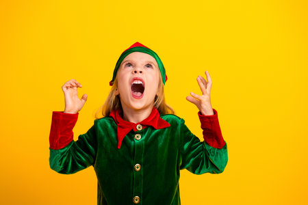 Excited young girl dressed as a festive elf yelling with enthusiasm against a vibrant yellow background during Christmas seasonの写真素材