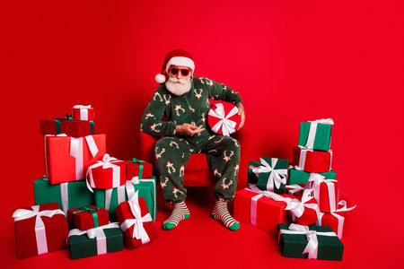 Santa in cozy pajamas and sunglass sits among a pile of colorful wrapped gifts in a bright red studio for Christmas Eveの写真素材