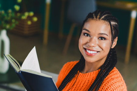 Bright smiling woman in orange sweater reads a book at home in a cozy interior by the window