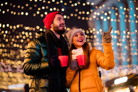 Fun loving couple in winter city lights enjoying hot drinks under festive decor and garlands as...
