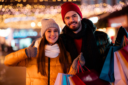 Two happy friends in warm coats pose with colorful shopping bags under festive lights enjoying Christmas shopping in the city at night
