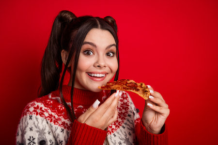 Smiling young woman in a red Christmas sweater enjoying a slice of pizza against a bright festive red backgroundの写真素材