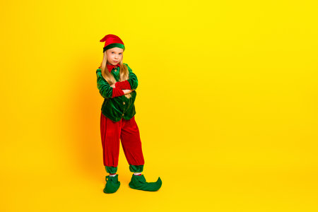 Young girl in a festive elf costume crossing arms playfully on a vibrant yellow background showcasing holiday cheerの写真素材