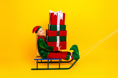 Adorable little elf in festive clothes sitting with stacked gifts on sled in front of a vibrant yellow backgroundの写真素材
