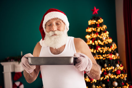 Santa Claus in a casual singlet baking in front of a decorated Christmas tree, spreading holiday joy and festive spiritの写真素材