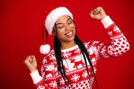 Playful young woman in Santa hat and red reindeer sweater celebrates Christmas joy against a bright red backdropの写真素材