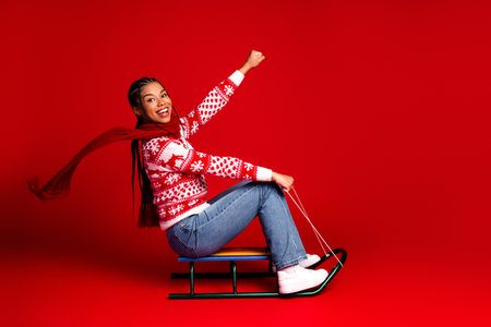 Playful seasonal portrait of a young woman on a sled in a red holiday backdrop celebrating winter joy and Christmas cheerの写真素材