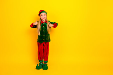 Little girl dressed as an elf against a vibrant yellow background showing cheerful thumbs up during...